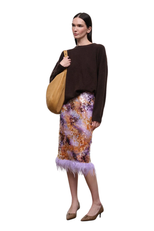 A model stands against a white background wearing a dark brown knit sweater, a purple and orange patterned feather-trimmed pencil skirt, and brown pointed-toe heels, while carrying a tan shoulder bag.