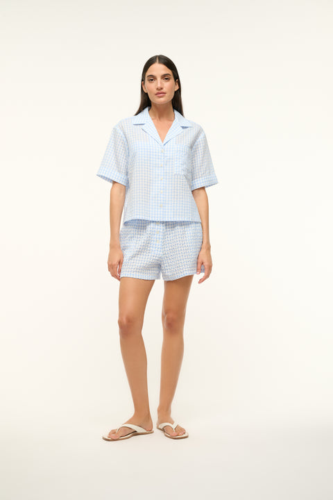 Woman wearing a blue gingham, short-sleeve, button up shirt with a pair of matching shorts