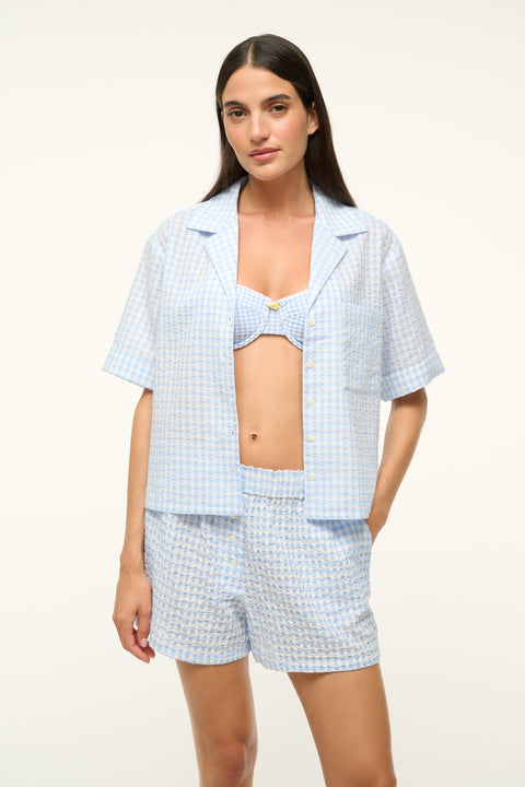 Close up of a woman wearing a blue gingham, short-sleeve, button up shirt undone with a pair of matching shorts