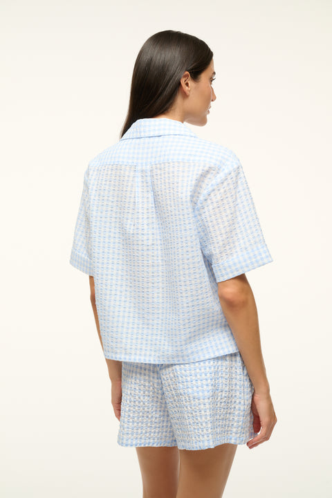 Back view of a woman wearing a blue gingham, short-sleeve, button up shirt with matching shorts