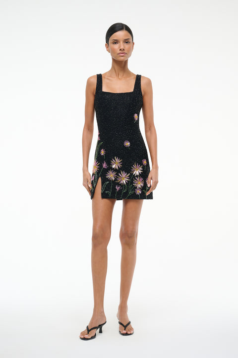 Woman wearing a sleeveless beaded mini dress that features a square neck and side slit, designed with pink dahlias on a black background, paired with black heels. 