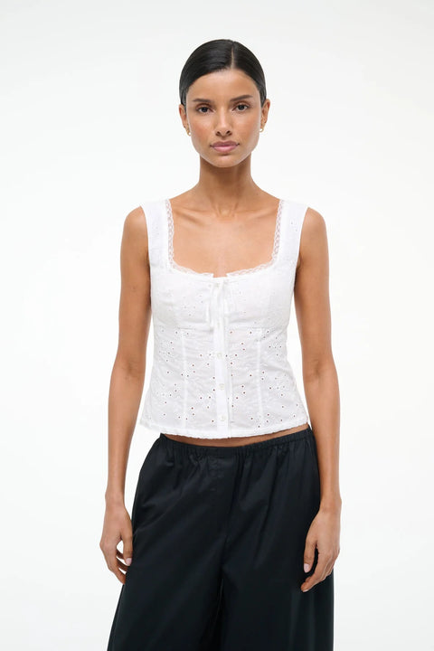 Woman wearing a white eyelet tank top that features lace trim, a button up detail and a ribbon tie at the neckline, paired with black pants. 