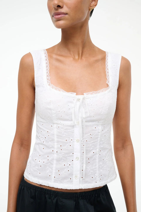 Close up of a woman wearing a white eyelet tank top that features lace trim, a ribbon tie neckline, and a button up detail. 
