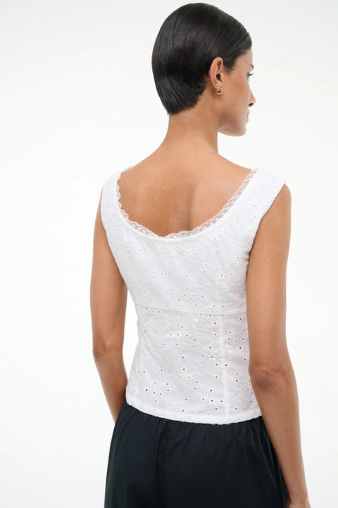 Back angle of a woman wearing a white eyelet tank top that features white lace trim and a scoop back, paired with black pants. 