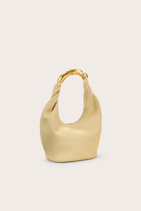 Side view of a pale yellow, leather top handle bag that features an abstract go9ld-toned handle. 