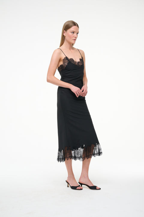 Side-facing woman wearing a black midi slip dress that features black lace trim on the neckline and hem, paired with black kitten heels. 