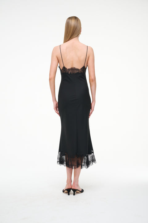 back-facing woman wearing a black midi slip dress that features black lace trim on the neckline and hem, paired with black kitten heels. 