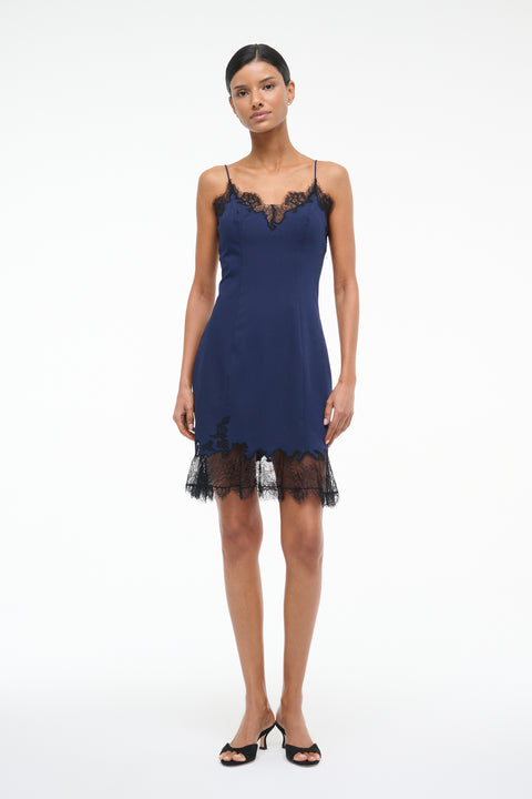 Woman wearing a navy blue mini slip dress with black lace trim on the neckline and hem, paired with black kitten heels.