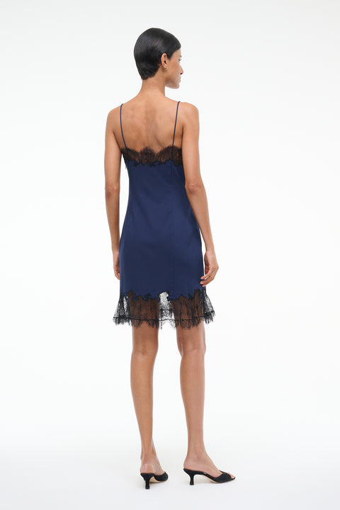 Back-facing woman wearing a navy blue mini slip dress with black lace trim on the neckline and hem, paired with black kitten heels.