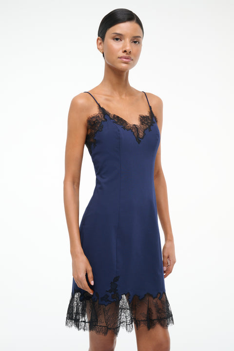 Close up of a woman wearing a navy blue mini slip dress with black lace trim on the neckline and hem. 