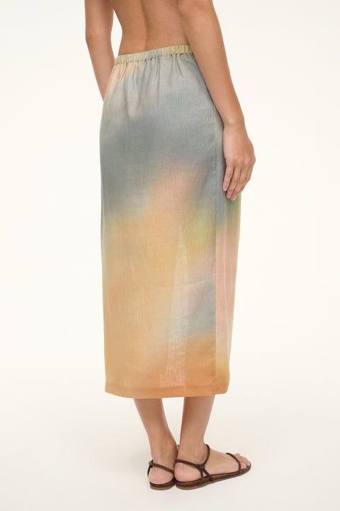 Back view of a woman wearing a pastel multicolor linen midi skirt that features an elastic waistband. 