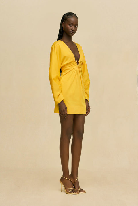 Woman wearing a yellow dress with a cinched ring under the bust against a beige background from the side view.