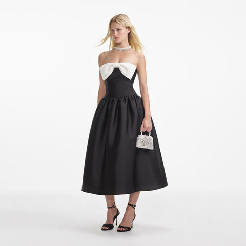 Woman in the black midi dress with white bow detail holding a handbag.