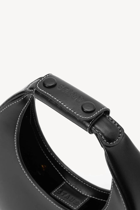 Detail shot of a small, black leather, moon shaped handbag with white stitching and a black leather strap on the handle. 