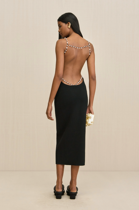 Woman wearing a black midi dress that features an open back with delicate straps lined with abstract pearls lining the hips and shoulders, paired with black sandals. 