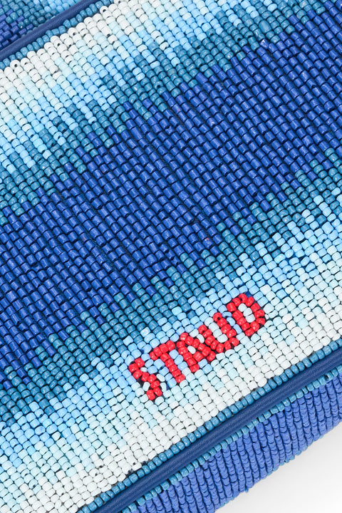 Detail shot of a glass beaded shoulder bag that features dark blue to white ombre stripes in a subtle wave pattern with STAUD written in red on the back. 