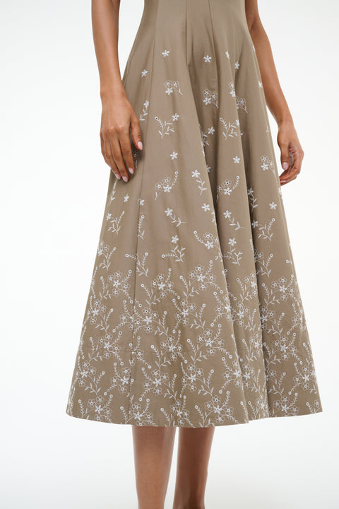 Detail shot of a woman wearing a taupe midi dress that features a fitted bodice and A-line skirt with white floral embroidery. 