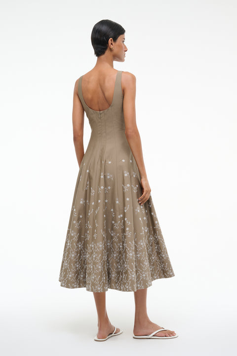 Back-facing woman wearing a sleeveless taupe dress that features a scoop back, fitted bodice, and A-line skirt with white floral embroidery, paired with white sandals. 