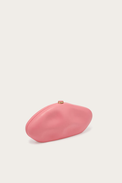 Side view of an abstract, oblong clutch covered in smooth pink leather that features indents for a sculptural look, complete with gold-toned hardware. 