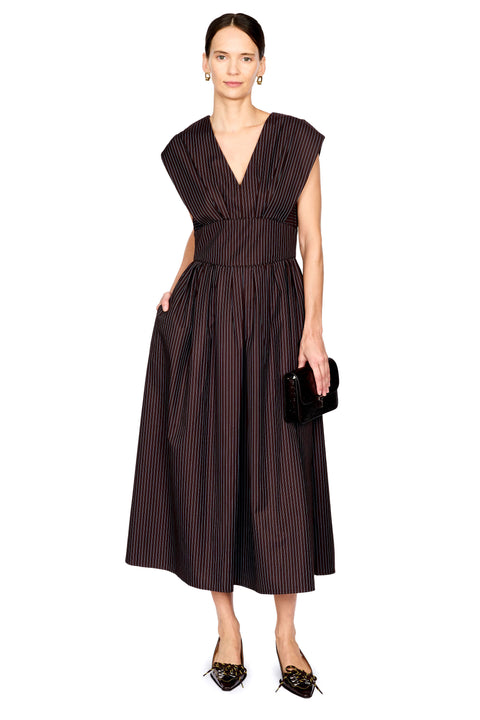 A woman modeling a sleeveless dark brown midi dress with thin vertical pinstripes, a V-neckline, and a gathered waist, paired with black pointed shoes and a small black clutch, against a plain white background.