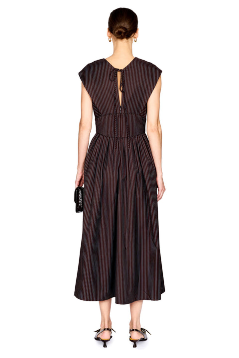 A woman modeling a sleeveless dark brown midi dress with thin vertical pinstripes, a V-neckline, and a gathered waist, paired with black pointed shoes and a small black clutch, against a plain white background.