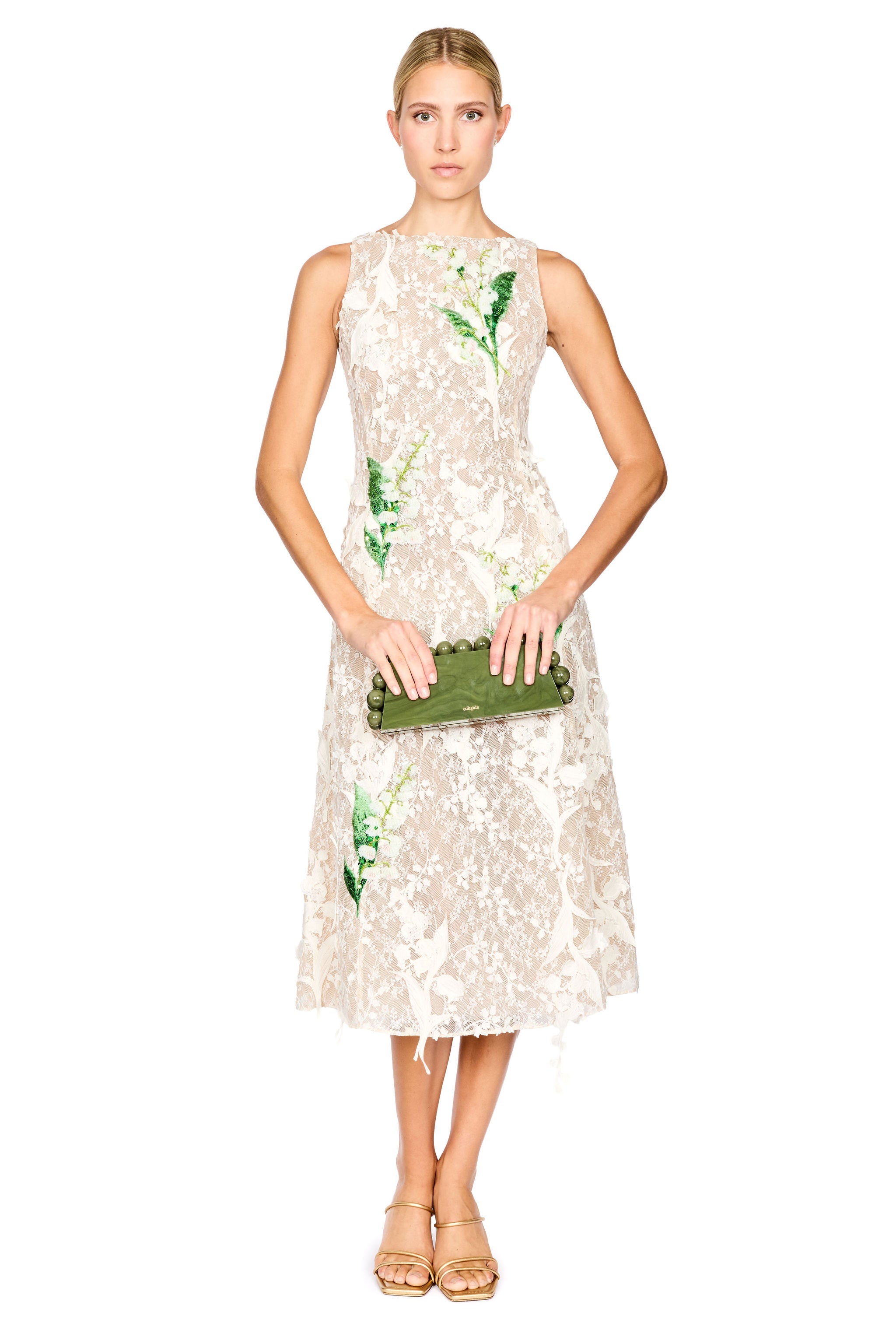 A model wearing a sleeveless cream lace midi dress with green floral applique, paired with gold high-heeled sandals, against a plain white background.