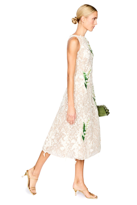 A model wearing a sleeveless cream lace midi dress with green floral applique, paired with gold high-heeled sandals, against a plain white background.