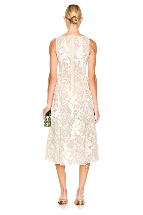 A woman viewed from the back wearing a sleeveless cream lace midi dress with a fitted bodice and flared skirt, holding a small green patterned clutch against a plain white background.