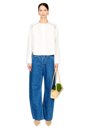 A model stands against a white studio background wearing a white long-sleeve blouse with decorative studded details on the shoulders, blue wide-leg jeans, and holding a beige tote bag.