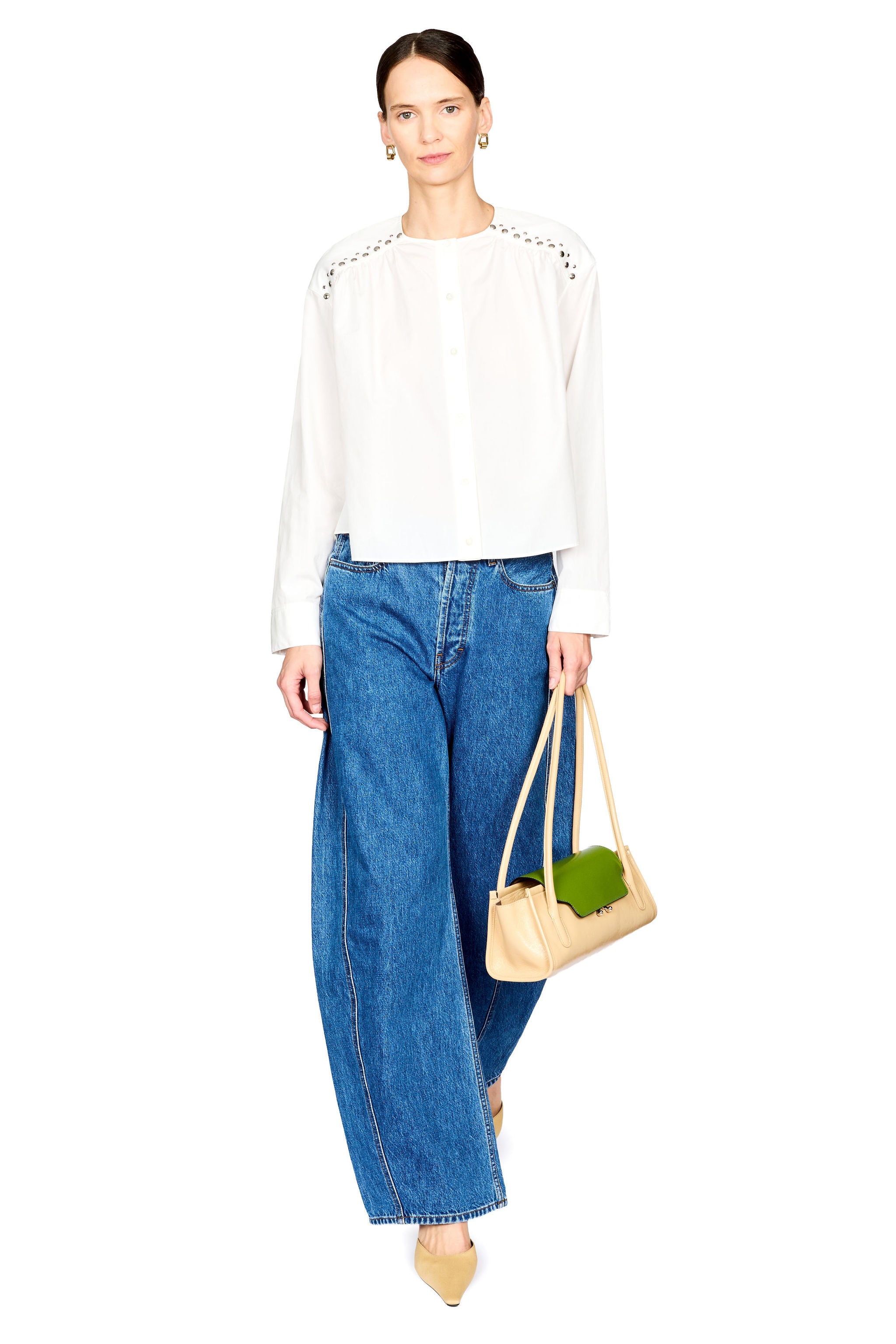 A model stands against a white studio background wearing a white long-sleeve blouse with decorative studded details on the shoulders, blue wide-leg jeans, and holding a beige tote bag.