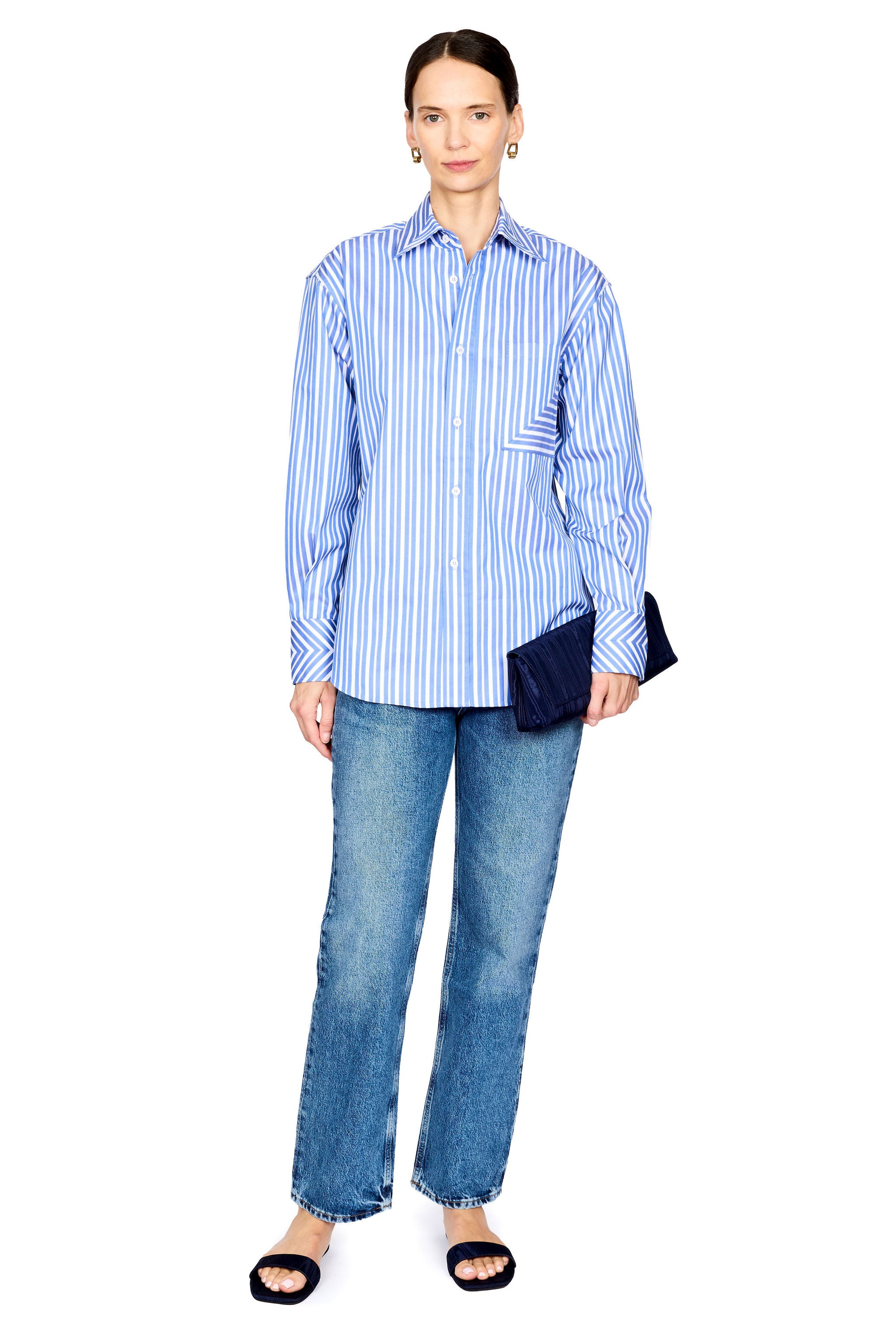 A woman modeling a light blue and white vertically striped button-down shirt with blue jeans, black slide sandals, and a dark navy clutch, set against a plain white background.