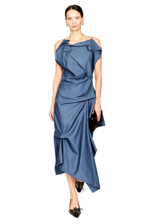 A woman modeling a blue one-shoulder midi dress with a draped, ruffled design and an asymmetrical hem, paired with black pointed flats and a small black clutch, against a plain white background.