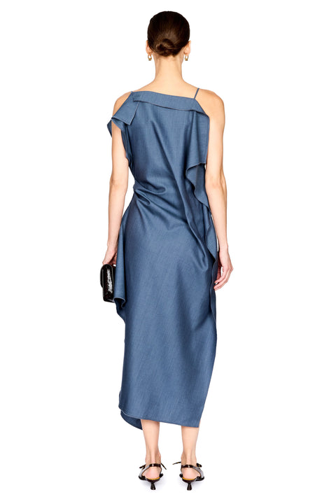 A woman viewed from the back wearing a slate blue one-shoulder midi dress with draped ruffle details, paired with black strappy heels and holding a small black clutch, against a plain white background.