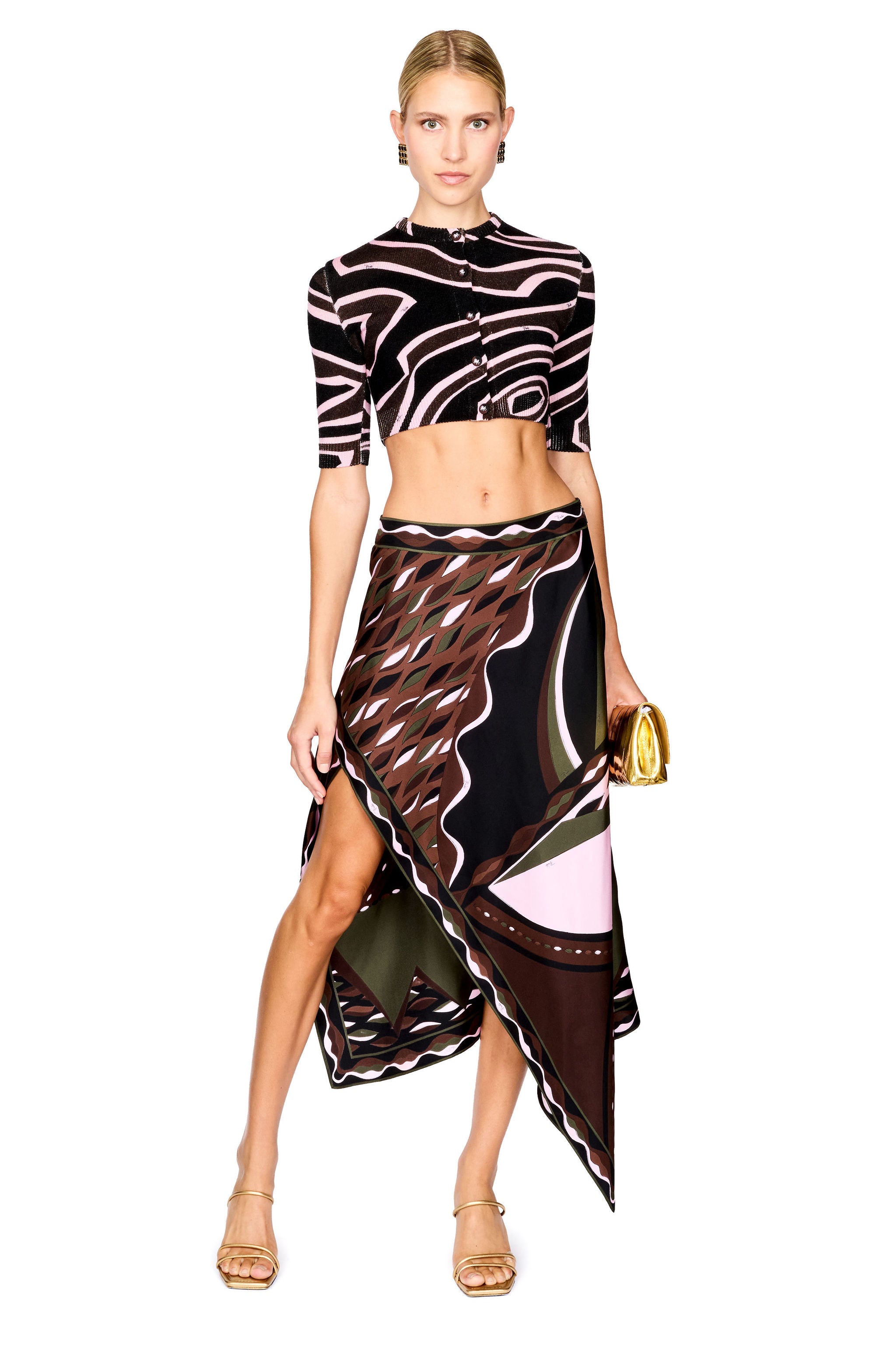 A model stands against a white studio background wearing a matching two-piece set with a black, pink, and brown wavy abstract print on a short-sleeve crop top and a high-low wrap skirt with pink and olive green accents. She is wearing gold strappy heels and holding a small gold clutch.