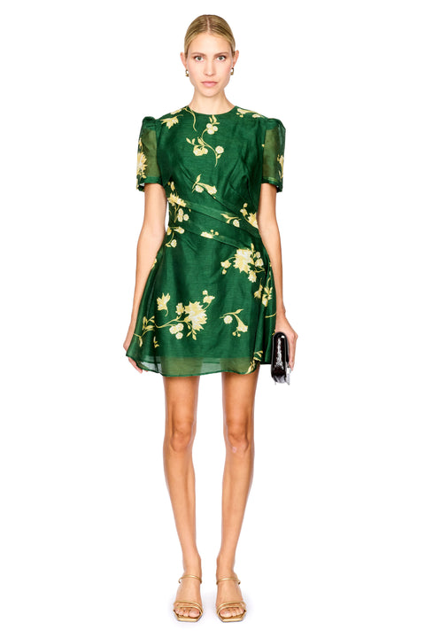 A model wearing a short-sleeved green dress with a yellow floral print, holding a small black clutch, set against a plain white background.
