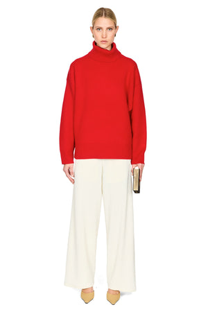 A model stands against a plain white background wearing a bright red turtleneck sweater and off-white wide-leg trousers, holding a slim black clutch.