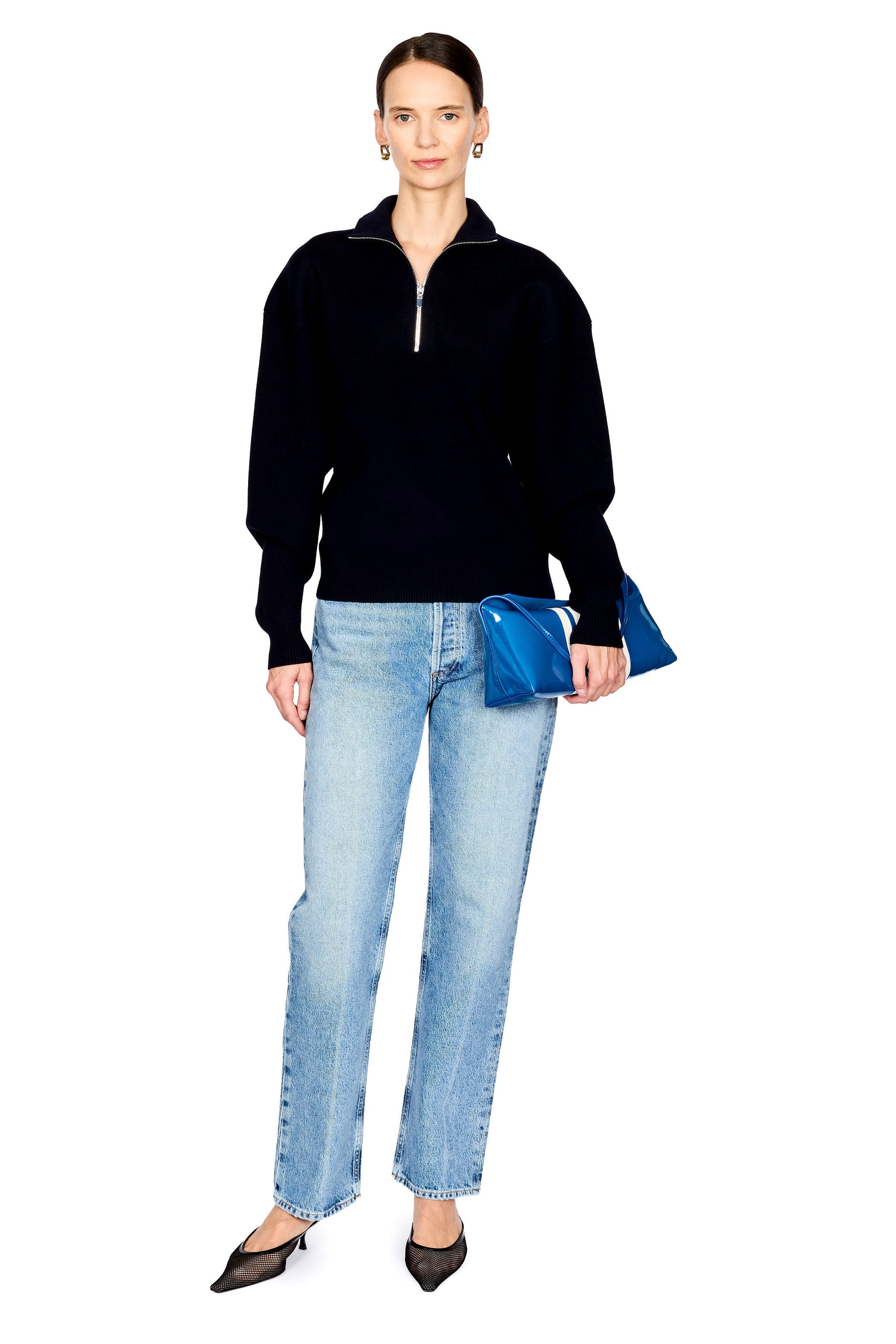 A woman modeling a navy long-sleeve quarter-zip sweater, light-wash blue jeans, and black pointed shoes, while holding a shiny blue handbag with white stripes against a plain white background.