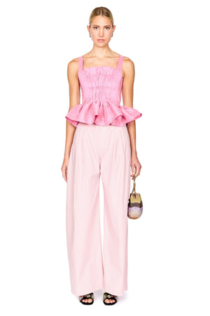A model stands against a white background wearing a light pink two-piece outfit consisting of a smocked sleeveless top with a ruffled hem and matching wide-leg trousers. She is holding a small handbag.
