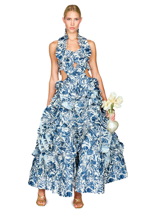 A model wearing a sleeveless, tiered halter maxi dress with a blue and white tropical floral print, holding a small vase of white flowers against a solid white background.