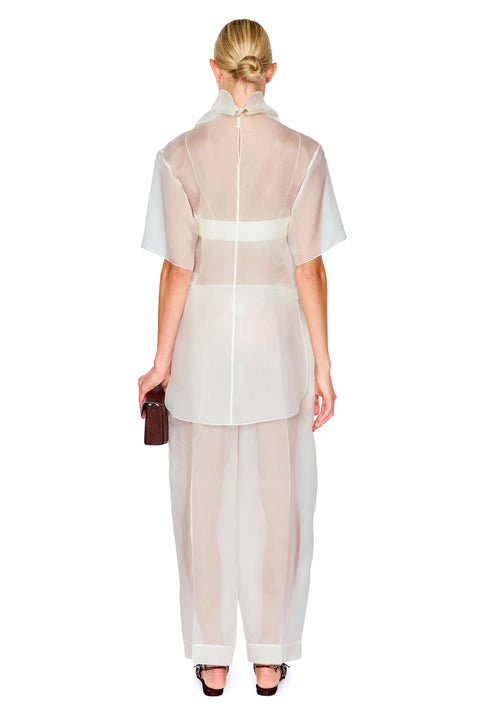 A model shown from the back wearing a sheer, semi-transparent white outfit consisting of a short-sleeve top and matching wide-leg pants, paired with black sandals and holding a small dark brown clutch against a solid white background.