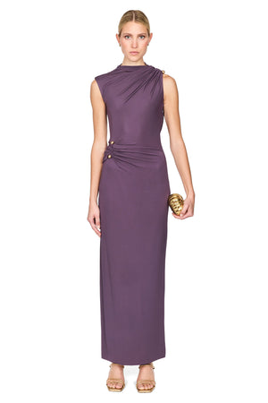 A woman modeling a sleeveless, high-neck, floor-length mauve dress with draped detailing and gold buttons, standing against a plain white background.