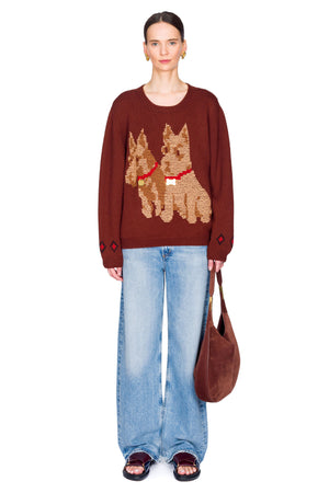 Person wearing a brown sweater with dog designs, blue jeans, and carrying a brown bag on a white background.