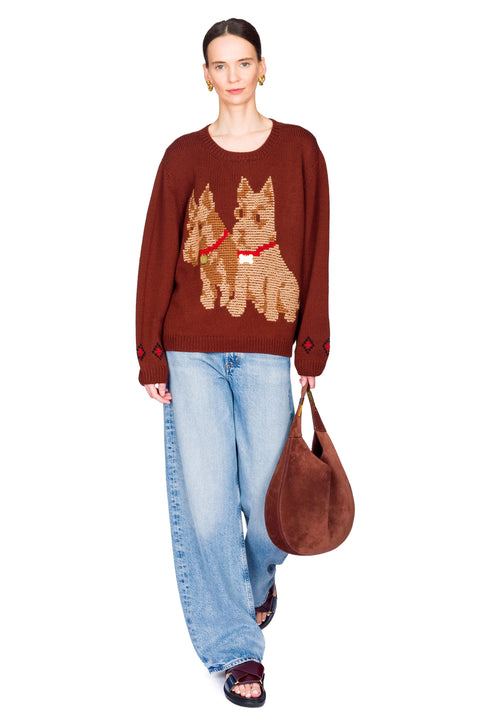 Person wearing a brown sweater with dog designs, blue jeans, and carrying a brown bag on a white background with one hand clutching bag.
