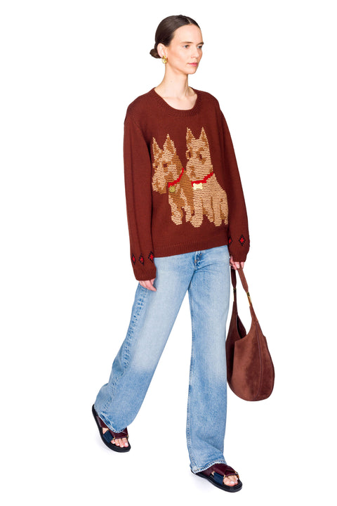 Person wearing a brown sweater with dog designs, blue jeans, and carrying a brown bag on a white background from side view.