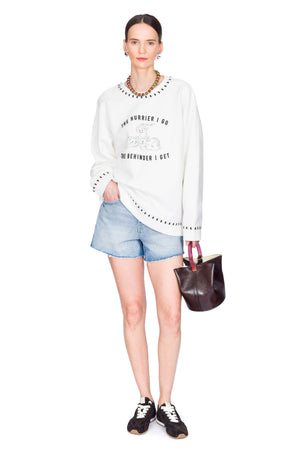 Woman wearing a white sweater with text, denim shorts, and holding a brown bag on a white background.