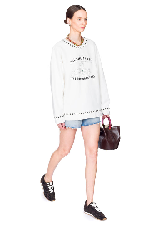 Woman wearing a white sweater with text, denim shorts, and holding a brown bag on a white background from the side.