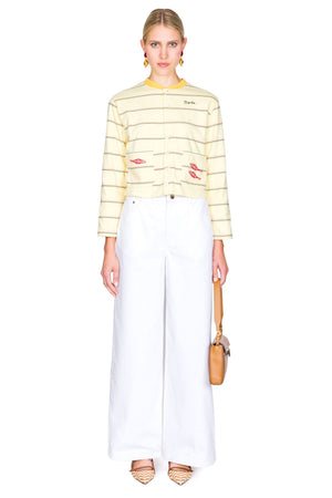 Woman wearing a yellow striped sweater with red fish on the pockets and white pants on a white background with one hand clutching bag.