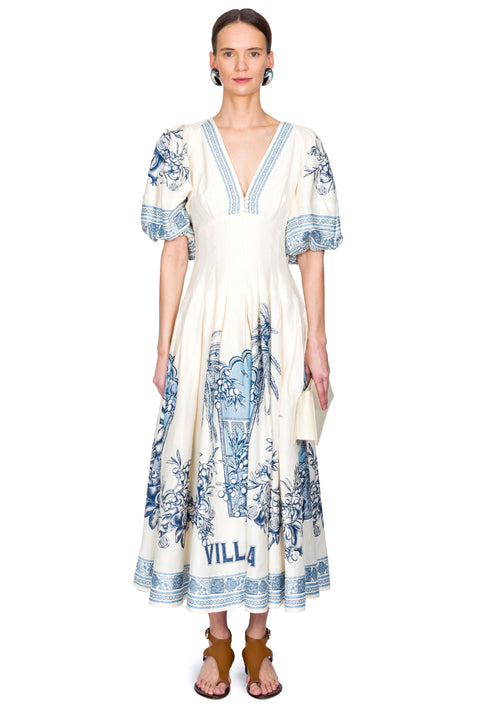 Woman wearing a white maxi dress with blue floral patterns and 'Villa' branding on a white background