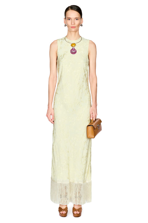 A model wearing a sleeveless, pale yellow maxi dress with a textured pattern and a fringed hem, accessorized with a colorful pendant necklace and a brown leather clutch bag, set against a plain white background.
