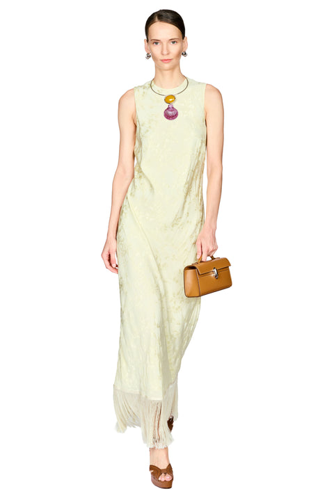 A model wearing a sleeveless, pale yellow-green maxi dress with a textured pattern and a fringed hem, accessorized with a colorful pendant necklace and a brown leather handbag, set against a plain white background.